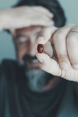 Migren ağrılarına karşı kırmızı hap tutan adama yaklaş. Arka plandaki kişi acıyla ön tarafına dokunuyor. Eczane ve ilaç tedavisi kavramı. İlaçlarla eline odaklan.