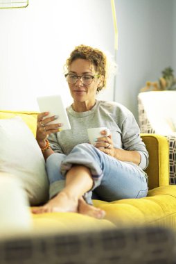 Modern orta yaşlı kadın tablet üzerinde kitap okuyor. Evdeki kanepede rahat rahat oturuyor ve evde tek başına vakit geçiriyor. Kafkasyalı kadın dijital cihazda çalışıyor.