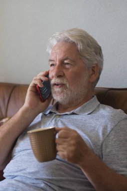 Evde modern telefonla konuşan, kahve içen ya da çay içen ve teknoloji bağlantısının tadını çıkaran yaşlı bir adam. Yetişkin bir erkek cep telefonunu evde dinlenirken ve eğlenirken kullanıyor.