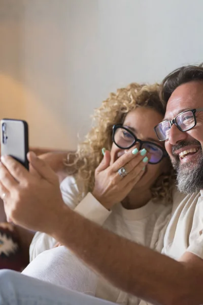 Happy  Adult Couple Making Video Call On Phone - cheerful people speak and listen friends online with cellular from home sitting on the sofa - holidays remote celebration concept