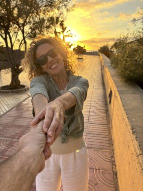 Adult couple walking with joy in sunset time holding hands. Man pov and woman smiling. People enjoying outdoor leisure activity together and summer holiday vacation. Travel and tourist concept lifestyle