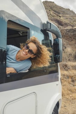 Mutlu ve özgür bekar bir kadın. Kamp arabasının penceresinin önündeki varış yerinin keyfini çıkarıyor ve gülümsüyor. Tatilde seyahat etmek için karavan kiralama fikri. Minibüs hayatı ve şebeke dışı