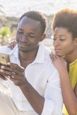 Genç siyah çift açık havada modern bir telefon kullanıyor. Güzel insanlar. Erkek ve kadın, arkadaşlarıyla birlikte yaşam tarzı teknolojisinden ve mesajdan zevk alıyor.