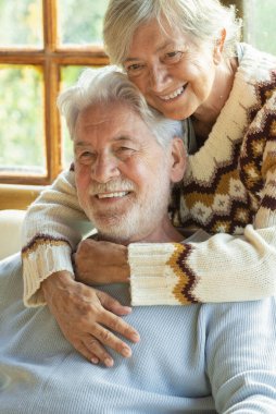 Happy and enjoyed senior caucasian couple smile and hug each other sitting on a chair in cozy home cabin chalet - two man and woman old elderly people lifestyle enjoying relax and relationship