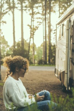 Doğanın tadını çıkaran sakin bir kadın arka planda yüksek ağaçlar ve ormanlar olan modern bir karavan karavanın önünde oturuyor. Gün batımı güneş ışığı. İnsanlar ve seyahat. Olay yeri otopark aracı. Yaşam.