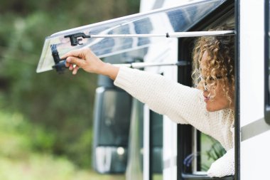 Arka planda yeşil dağ ormanı olan bir kadın karavan penceresi açıyor. Seyahat ve gezgin kavramı. Araç seyahati tatil yaşam tarzı. İnsanlar ve doğa. Vanlife tarzı. Özgürlük hayatı.