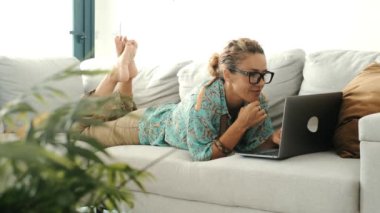  Modern kadın kanepede uzanıyor ve internette veya işte dizüstü bilgisayar kullanıyor. İnsanlar ev içi eğlence aktivitelerinde zaman ve teknoloji bağlantısından keyif alıyorlar.. 