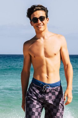 Mayo giymiş genç bir delikanlı arka planda mavi gökyüzü ve okyanus suyuyla sahilde yürüyor yaz tatilinin tadını çıkarıyor gülümsüyor ve kameraya bakıyor.