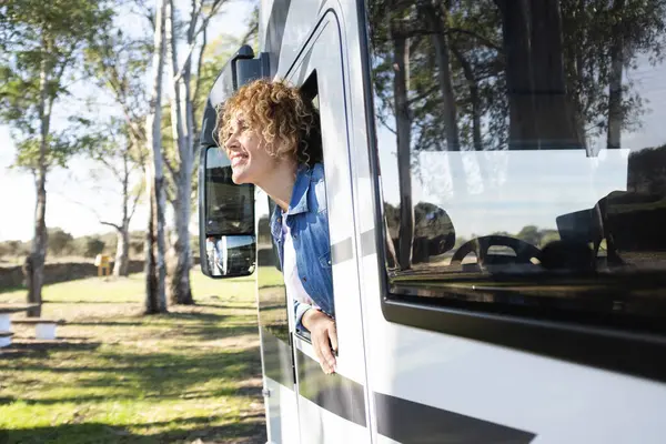 Beautiful smiling Caucasian curly woman leaning out of the window of a camper. Travel, nature and leisure concept