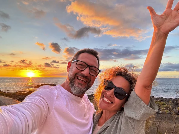 Happy adult couple in love taking selfie picture with romantic wonderful sunset on the ocean in background. Young mature people man and woman enjoying travel destination in summer holiday vacation