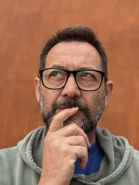 Pensive adult man lookig up and touching chin in outdoor leisure against a brown background. One male people wearing glasses with thoughts expression on face. Problems and life.