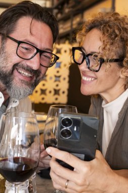 Lokantada gülümseyen ve konuşan mutlu çift cep telefonuna bakıyor, evli çift kafeteryada öğle yemeği yiyor, insanlar ve teknoloji kavramı.