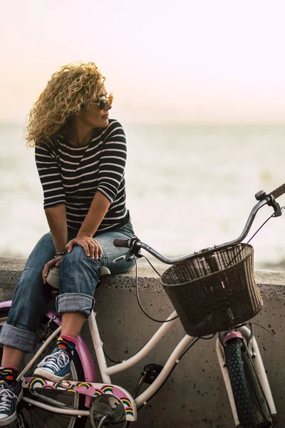 People enjoying outdoor leisure activity looking a beautiful colored sunset - beautiful adult young woman with long curly blonde hair  enjoy the relax after a bike tour - horizon and sun with ocean