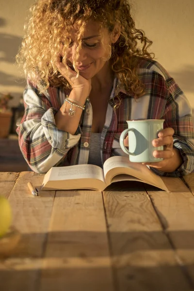Beautiful curly adult woman read a book outdoor at home in terrace or trendy room with wood table and tea or coffee - style lifestyle and concept of people enjoying life at home