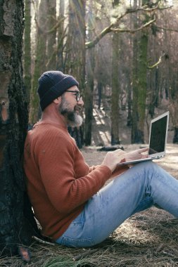  Orman odunlarında dizüstü bilgisayar kullanan bir adam teknolojinin ve boş zamanların tadını çıkarıyor. Modern insan arka planda ağaçlarla hücresel olarak konuşuyor. Seyahatte insanlar ve iletişim