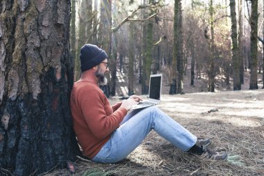  Orman odunlarında dizüstü bilgisayar kullanan bir adam teknolojinin ve boş zamanların tadını çıkarıyor. Modern insan arka planda ağaçlarla hücresel olarak konuşuyor. Seyahatte insanlar ve iletişim