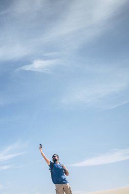 Açık havada duran adam boş zamanlarında internet sinyallerini kontrol ediyor. İnternet telefon aygıtı kavramı ve dünyadaki iletişim zamanı kavramını kontrol ediyor. Mavi gökyüzü geçmişi.