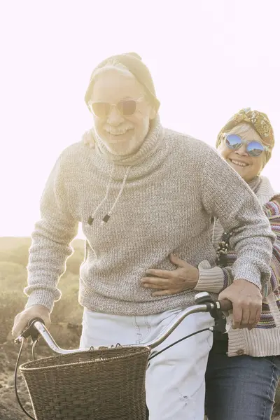 Couple of active cheerful and happy senior retired people enjoy together riding a bicycle in outdoor leisure activity with sun in backlight - concept of old age and fun joyful with no limit