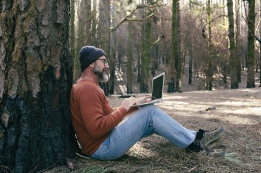  Orman odunlarında dizüstü bilgisayar kullanan bir adam teknolojinin ve boş zamanların tadını çıkarıyor. Modern insan arka planda ağaçlarla hücresel olarak konuşuyor. Seyahatte insanlar ve iletişim