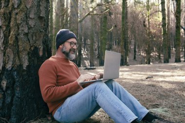  Orman odunlarında dizüstü bilgisayar kullanan bir adam teknolojinin ve boş zamanların tadını çıkarıyor. Modern insan arka planda ağaçlarla hücresel olarak konuşuyor. Seyahatte insanlar ve iletişim