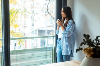 Kadın evde dikilip pencereden dışarı bakıyor bir fincan kahve içiyor ve gülümsüyor.. 