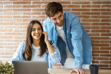 Genç mutlu çift gülümsüyor ve ev işyerindeki iş yerinin bilgisayarında birlikte bir dizüstü bilgisayar kullanıyorlar.. 