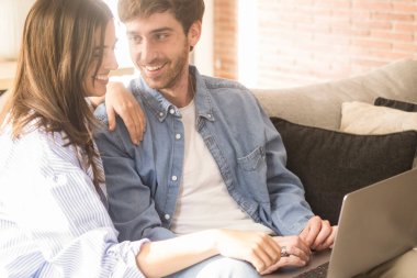 Evdeki kanepede birlikte dizüstü bilgisayar kullanan ve ekrana gülümseyen mutlu modern çift. Neşeli erkek ve kadın birlikte bilgisayar aktivitelerinin keyfini çıkarıyorlar. Bir sonraki tatil için otel rezervasyonu