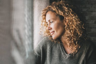Yetişkin bir kadın, yüzünde barışçıl bir ifade ve küçük bir gülümsemeyle pencereden dışarı bakıyor. Kapalı alandaki kendine güvenen kadınlar boş zamanlarını rahat geçirirler. Hayal kuran ve düşünen bayan