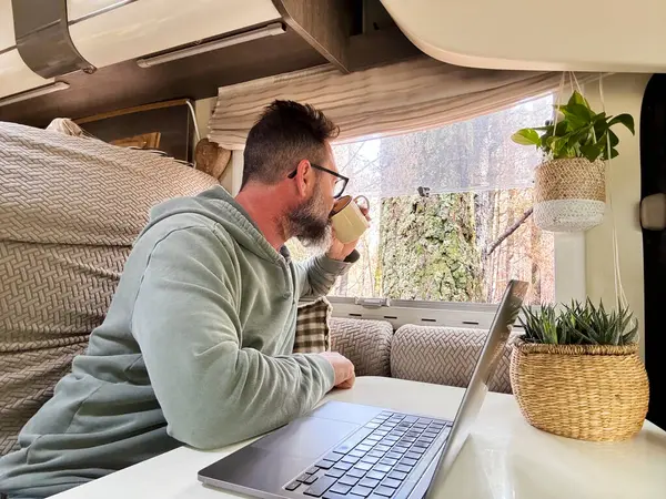 man enjoying camper van parking in the forest woods and admire nature outdoors park with coffee and laptop. Traveler people with motorhome. Nomadic vacation lifestyle. Mature male relaxing in indoor leisure activity