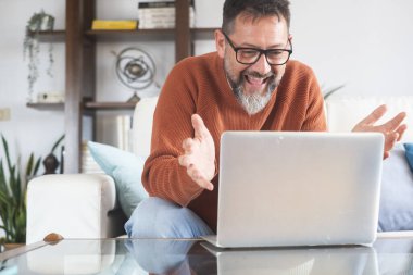Gözlüklü yetişkin bir beyaz adam evdeki koltukta oturup bilgisayar başında konuşuyor. İnternette çalışan serbest bir erkek, uzaktan kumandalı bir müşterisiyle internet üzerinden sanal konferans görüşmesi yapıyor.