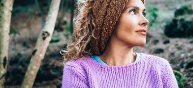 Çekici yetişkin bir kadının portresi, arka planda ağaçlarla birlikte doğa parkında hissetmenin keyfini çıkarıyor. Gezgin ve refah, doğal yaşam tarzı insan kavramı. Gülümseyen sakin kadın başka tarafa bakıyor.