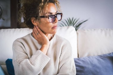 Kanepede yalnız başına oturan bir kadın boynuna dokunuyor ve masaj yapıyor, acı veriyor. Düşünceli kadın insanlar ve sağlıklı konsept. Çekici, düşünceli bir bayan depresif bir ifadeyle dışarı bakıyor.