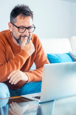 Yetişkin serbest çalışan bir adam dizüstü bilgisayara bakıp düşünüyor ve çenesine dokunuyor. Olgun erkek gözlüğü takıyor. Uzaktan kumandalı iş konsepti yaşam tarzında çalışan akıllı ev ödevleri. Kanepede oturan modern işçi.