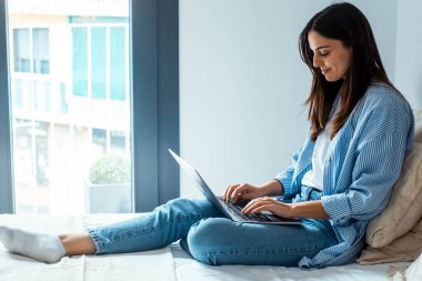 Genç bayan evde tek başına internette sörf yapıyor. Yatak odasında yatakta oturuyor.