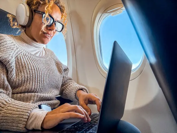 イヤホンで音楽を聴く快適な座席に座って ボード上のワイヤレス接続を使用して現代のノートパソコンで作業している魅力的なブロンドの乗客 — ストック写真