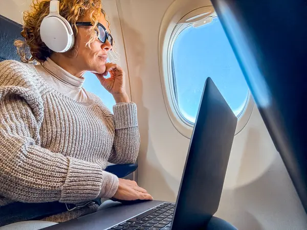 イヤホンで音楽を聴く快適な座席に座って ボード上のワイヤレス接続を使用して現代のノートパソコンで作業している魅力的なブロンドの乗客 ロイヤリティフリーのストック写真