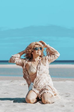Sahilde oturan kollarını açmış güzel bir kadının ön görüntüsü. Sarışın, kıvırcık saçlı, rüzgarda savrulan kadın rahatlar ve gülümser..