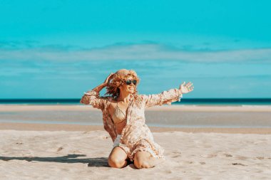Sahilde oturan kollarını açmış güzel bir kadının ön görüntüsü. Sarışın, kıvırcık saçlı, rüzgarda savrulan kadın rahatlar ve gülümser..