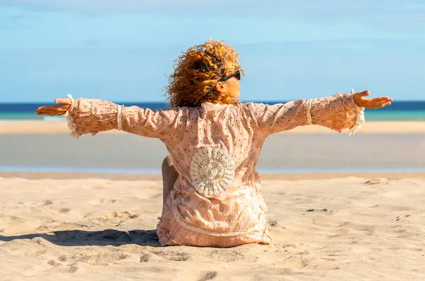 一个美丽的女人 张开双臂坐在海滩上的背影 金发女人 卷曲的头发在风中飘扬 放松地望着大海 微笑着 图库照片
