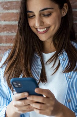 Genç, modern, beyaz kız gülümseyen telefona bakıyor, duvara yaslanmış duruyor. Rahatlamış bir kadın telefona bakar. Teknoloji konsepti.