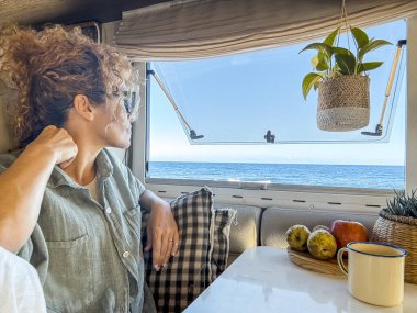 Gözlüklü kıvırcık kadın çay içerken gülümsüyor ve karavanının camından dışarı bakıyor. Evde tekerlekli sandalyede tatil yapar ve deniz kenarında bahar macerası yaşar. Göçebe yaşam tarzı konsepti