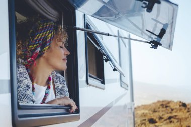 Kafasında atkısı olan kıvırcık saçlı bir kadınla kampçı kampın camına yaslanmış. Dağın eteklerinde turist durağı olan minibüs manzarayı daha iyi görebilmek için duruyor. Kamp seyahati ve özgürlük kavramı.