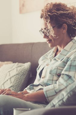 Rahat, olgun bir kadın kanepede gülümseyen dizüstü bilgisayar kullanıyor ve evde rahatça eğleniyor. İnternette internetten bağlanan mutlu kadın insanlar. Modern ev yaşam tarzı