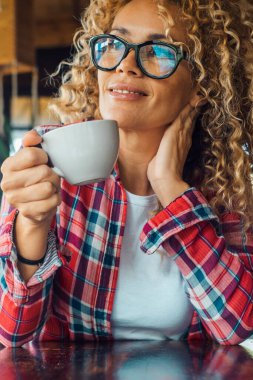 Serene woman at home drinking tea or coffee and enjoying relax leisure indoor activity alone sitting at the table. Portrait of cheerful and happy female people smiling and wearing eyewear. Curly hair