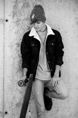 Şehir tarzı giysiler içinde poz veren ve beton duvara karşı uzun paten kayan genç bir gencin tam boy portresi. Siyah-beyaz fotoğrafçılık modası