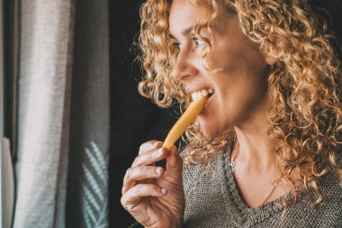 Uzun kıvırcık saçlı, sağlıklı meyve yiyen ve pencereden dışarı bakan kadının yan portresi. Çekici kadın sağlığı iyi doğal yaşam tarzı yer. İçeriyi düşünüyorum. Gülümseyen kadın