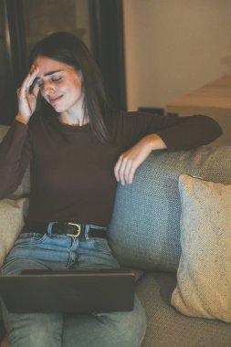 Evdeki kanepede oturmuş ağır baş ağrısı olan dizüstü bilgisayarı kullanan stresli bir kız, bilgisayar başında çalışırken migren ağrısı çeken genç bir kadın, yorgun hisseden bir kadın.