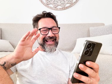 Yetişkin bir adam evde tek başına akıllı telefondan yapılan telefon görüşmesinin keyfini çıkarıyor. Elleriyle işaret ediyor ve mutlu bir şekilde gülümsüyor. Modern insanlar kapalı alanda boş zaman aktivitesi. Olgun erkek çevrimiçi arıyor