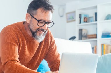  Yetişkin bir adam evde dizüstü bilgisayar kullanarak oturuyor gülümsüyor ve kablosuz teknolojinin ve bilgisayar bağlantısının tadını çıkarıyor. Küçük işletme ve alternatif ofis, modern insan yaşam tarzı. Erkek sakalı