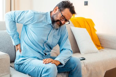 Adam kötü bir duruş için koltuğunun altına masaj yapıyor. İnsanlar evlerinde sağlıksız alışkanlıklar edinirler. Olgun erkek yüzüne acı veren bir ifadeyle vücuduna masaj yapar.